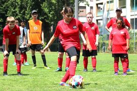 Eine Gruppe junger Fußballspieler in roten Trikots steht auf einer Wiese. Ein Spieler kickt einen bunten Fußball, während andere zuschauen, einige mit den Händen in den Hüften. Im Hintergrund sind Bäume und Gebäude zu sehen.