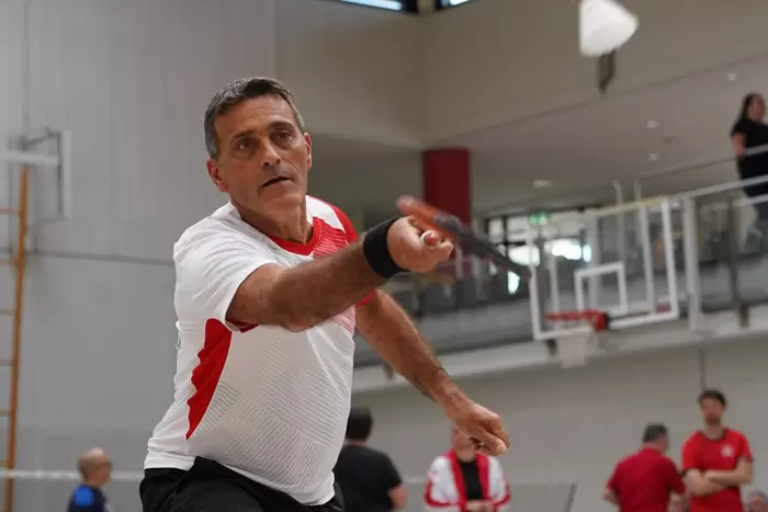Ein Mann in einem weiß-roten Hemd spielt in einer Halle Badminton, wobei er seinen Schläger nach vorne streckt, um einen Federball zu schlagen. Im Hintergrund sind andere Spieler und ein Basketballkorb zu sehen.