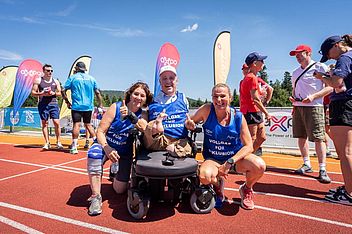 Drei lächelnde Sportler posieren auf einer Laufbahn, einer davon sitzt im Rollstuhl. Zwei weitere hocken daneben und zeigen den Daumen nach oben. Im Hintergrund sind unter einem strahlend blauen Himmel Menschen, Transparente und Fahnen zu sehen.