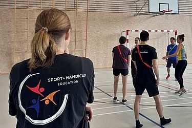 Eine Gruppe von Menschen in Sportkleidung steht in einer Sporthalle und hört sich Anweisungen an. Eine Person im Vordergrund trägt ein schwarzes T-Shirt mit dem Logo von „Sport Handicap Education“ auf dem Rücken.