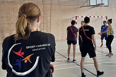 Eine Gruppe von Menschen in Sportkleidung steht in einer Sporthalle und hört sich Anweisungen an. Eine Person im Vordergrund trägt ein schwarzes T-Shirt mit dem Logo von „Sport Handicap Education“ auf dem Rücken.