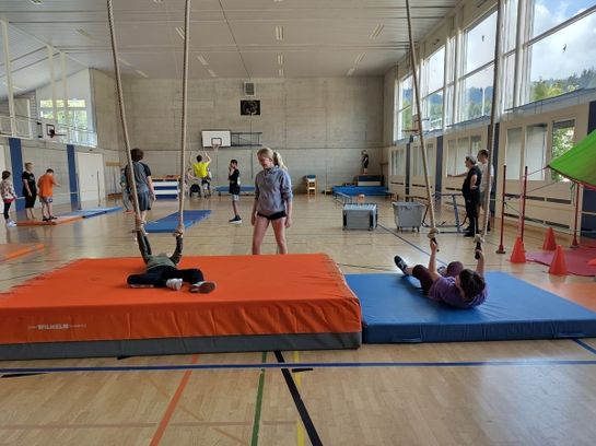 In einer geräumigen Turnhalle nehmen mehrere Kinder an Turnübungen teil. Zwei Kinder liegen auf Matten und halten Seile, während andere in der Nähe stehen, gehen oder warten. Sportgeräte und natürliches Licht erfüllen die Szene.