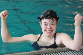 Ein Schwimmer mit Schwimmbrille auf dem Kopf lächelt freudig und hebt beide Arme zum Jubeln, während er in der Nähe des Randes eines Schwimmbeckens steht.