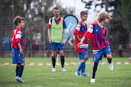 Vier Jungen in roten und blauen Fußballtrikots stehen auf einer Wiese, und einer von ihnen kickt einen Ball. Ein Erwachsener in einer grünen Weste steht hinter ihnen und beobachtet das Spiel. Im Hintergrund sind Bäume und ein Netz zu sehen.