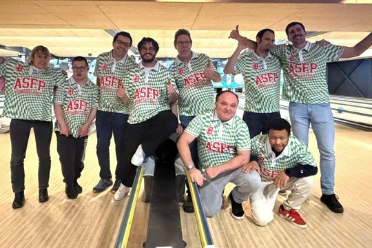 Eine Gruppe von neun Personen in passenden grün karierten ASFP-Hemden posiert und lächelt gemeinsam auf einer Bowlingbahn, einige stehend, andere sitzend, mit Bowling-Bahnen im Hintergrund.