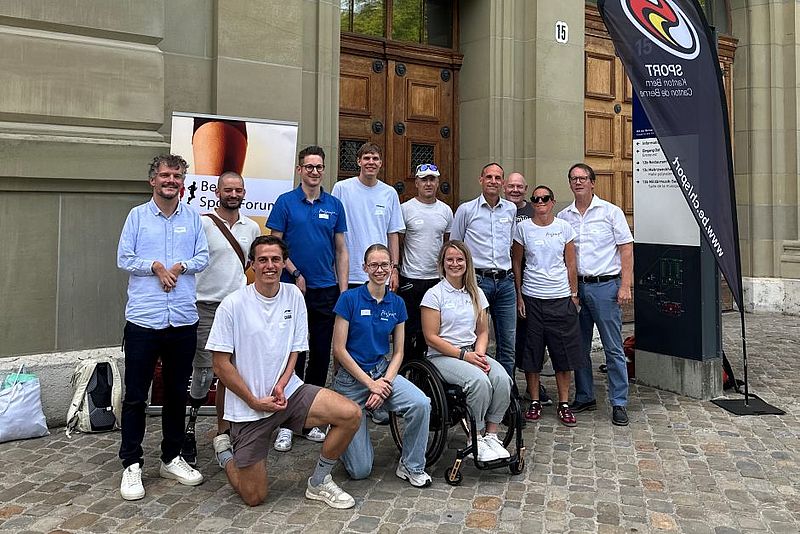 Eine Gruppe von Menschen, darunter eine Frau im Rollstuhl, posiert und lächelt gemeinsam vor einem Gebäudeeingang, neben Transparenten und einer Flagge auf einem gepflasterten Vorplatz.