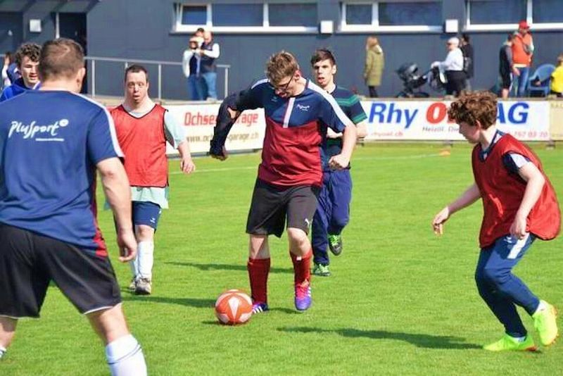Eine Gruppe junger Männer spielt Fußball auf einem Rasenplatz. Ein Spieler in einem roten Trikot dribbelt, während andere in blauen und roten Trikots versuchen, zu verteidigen oder zu unterstützen. Zuschauer beobachten die Szene von der Seitenlinie.