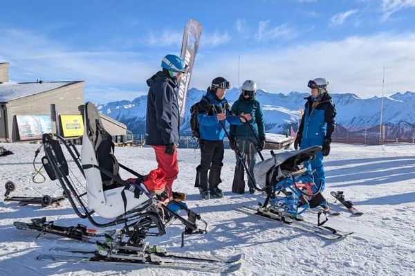 Mehrere Personen in Winterausrüstung unterhalten sich auf einem verschneiten Berghang in der Nähe von zwei Sitzskiern, mit schneebedeckten Gipfeln und einem klaren blauen Himmel im Hintergrund. Ein Gebäude und andere Skifahrer sind in der Nähe zu sehen.