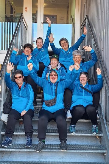 Sieben Frauen in passenden blauen Jacken sitzen und stehen auf einer Außentreppe, lächeln und heben feierlich ihre Arme. Der Schauplatz scheint sich vor einem Gebäude mit Glastüren zu befinden.