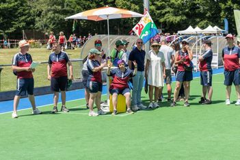 Eine Gruppe von Menschen in passenden Sportuniformen steht auf einer grünen Wiese, einige tragen Ausrüstung und eine bunte Fahne, während andere Hüte tragen und an einem sonnigen Tag unter einem großen Sonnenschirm stehen.