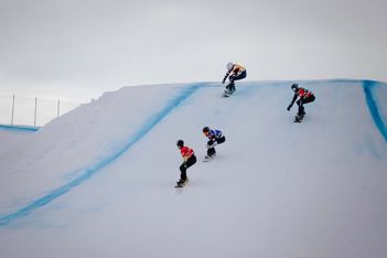 Vier Snowboarder in farbenfroher Ausrüstung rasen unter bewölktem Himmel eine verschneite, mit blauen Linien markierte Piste hinunter. Die Snowboarder fahren in verschiedenen Abständen und in unterschiedlichen Winkeln die Piste hinunter.