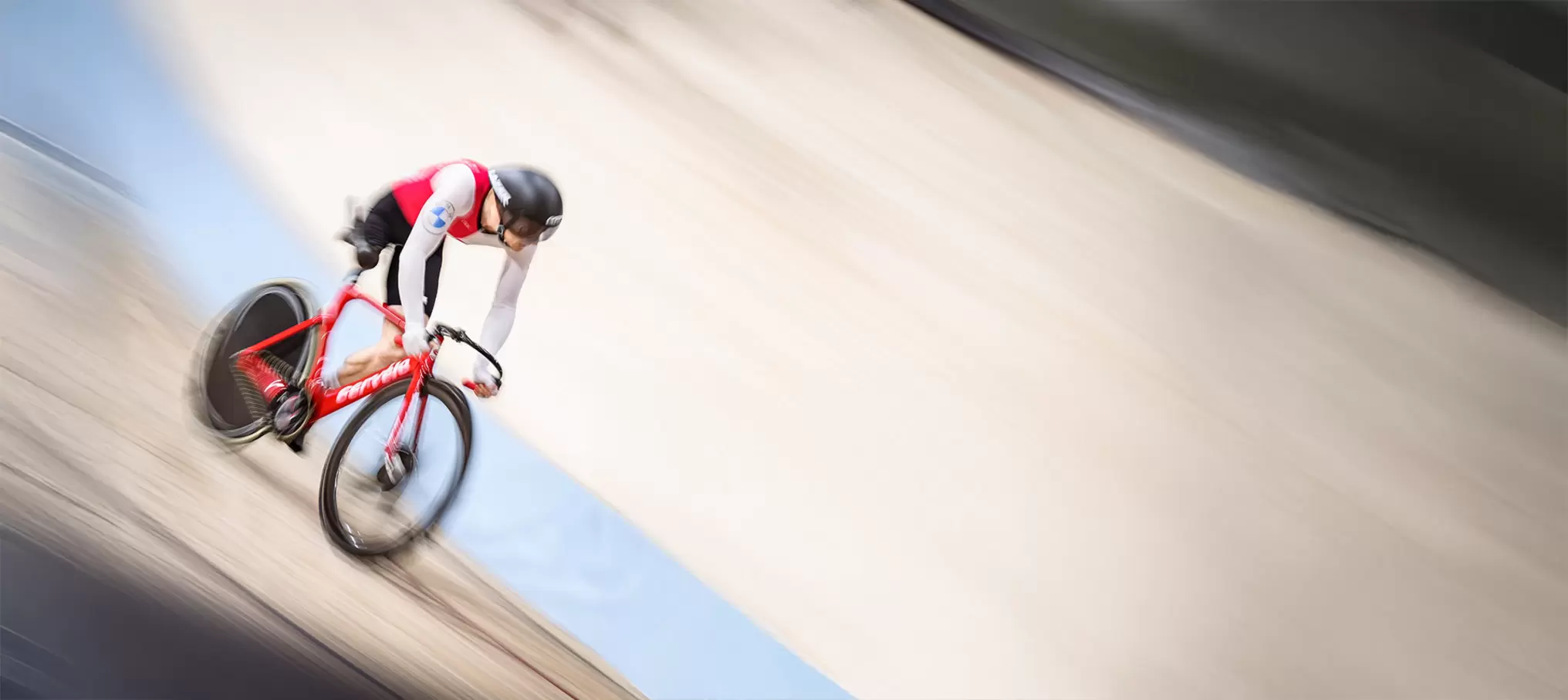 Ein Radfahrer in rot-weißem Outfit und schwarzem Helm rast über eine Velodrombahn, wobei der Hintergrund unscharf ist, um die Bewegung hervorzuheben.