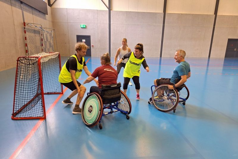 Vier Personen, zwei in Rollstühlen und zwei im Stehen, spielen in einer Turnhalle Rollstuhlhockey. Sie sind in der Nähe des Tores konzentriert und tragen gelbe oder blaue Sporttrikots, während im Hintergrund ein Netz und die Wände der Turnhalle zu sehen sind.