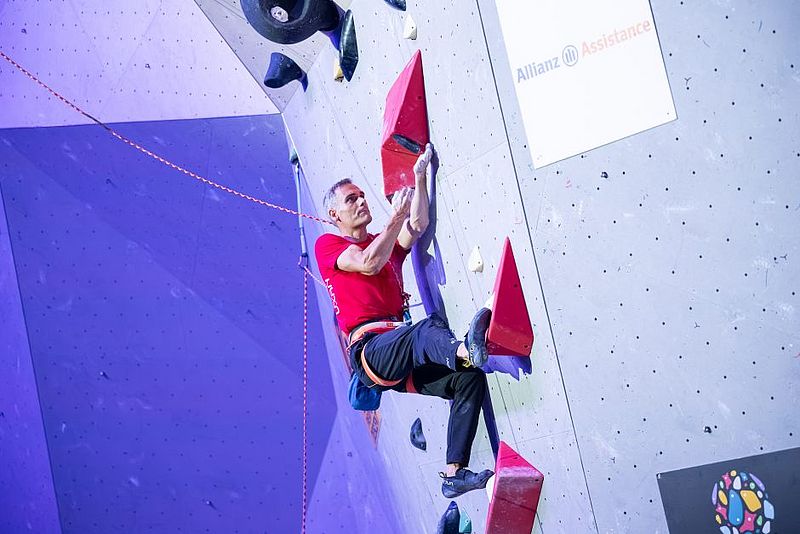 Ein Mann in einem roten Hemd und einer schwarzen Hose klettert an einer Indoor-Kletterwand mit roten und schwarzen Griffen, gesichert durch ein Seil und einen Klettergurt.