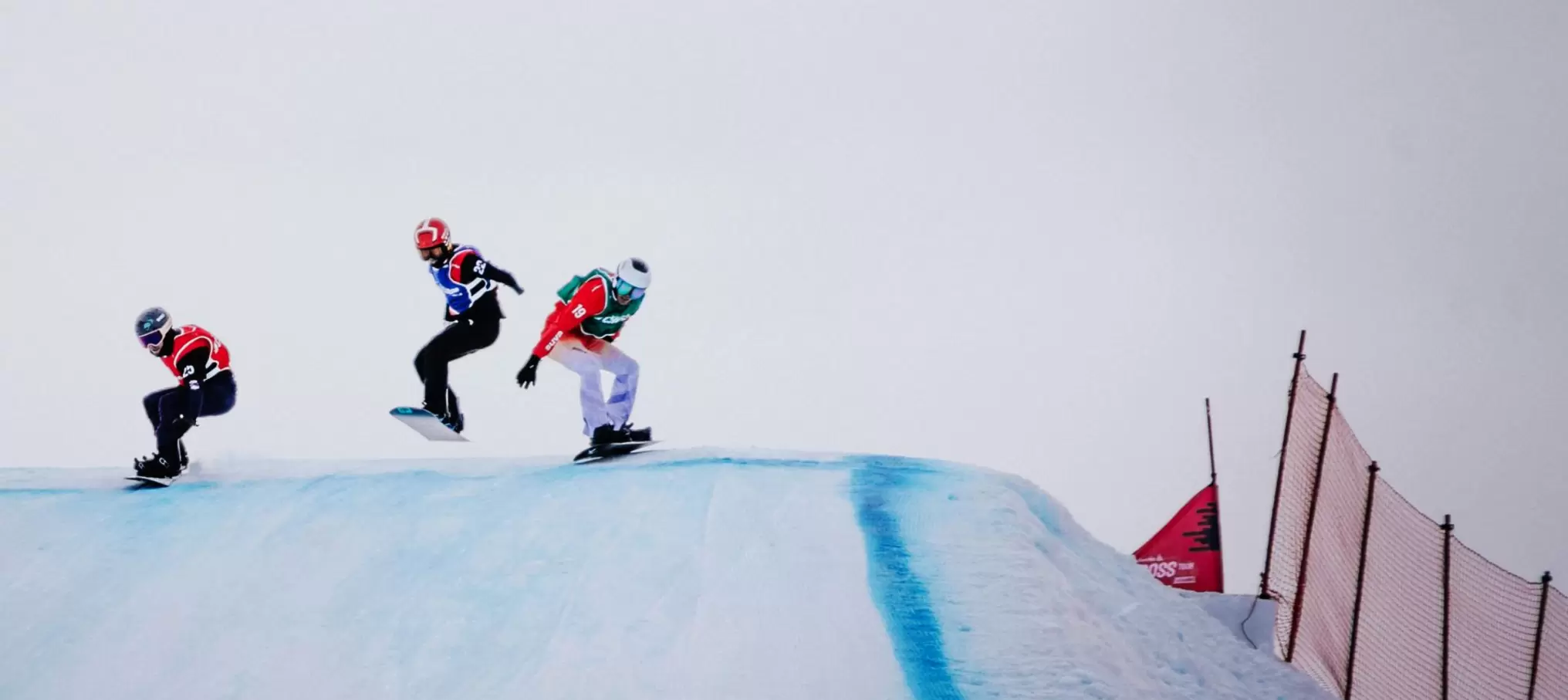 Drei Snowboarder mit Helmen und nummerierten Westen rasen eine verschneite Piste hinunter und fliegen über einen Grat. Rechts sind ein rotes Sicherheitsnetz und eine Flagge vor einem grauen Himmel zu sehen.