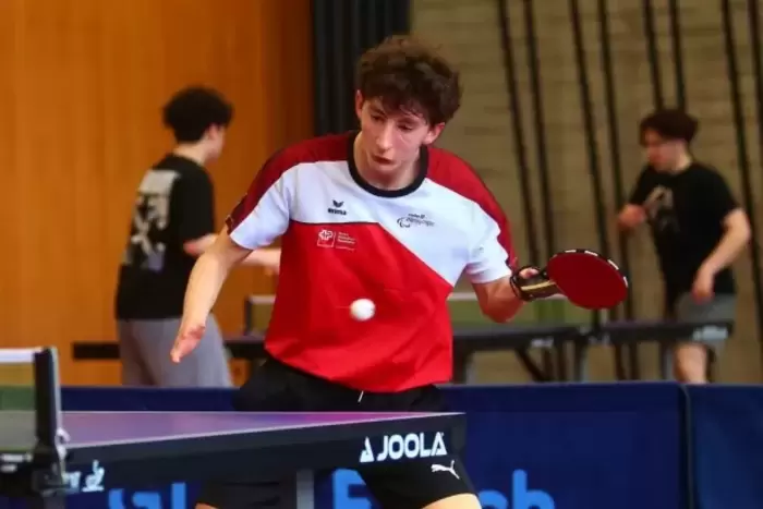 Ein junger männlicher Tischtennisspieler mit Beinprothese in Aktion, der ein rot-weißes Hemd und schwarze Shorts trägt und gerade einen Schlag abgibt. Zwei weitere Personen stehen im Hintergrund an einem anderen Tisch.