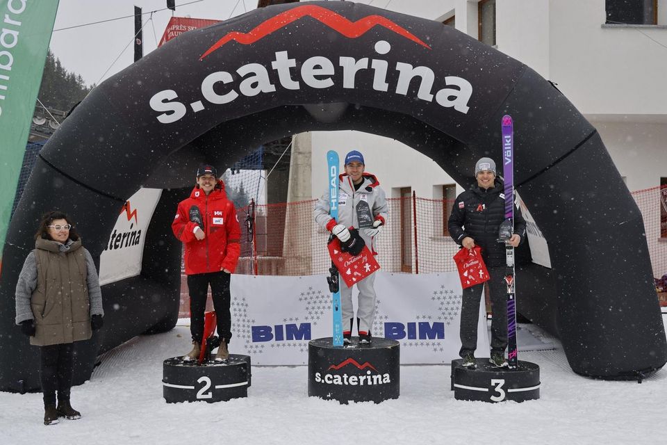 Drei Athleten stehen auf einem verschneiten Siegertreppchen unter einem schwarzen Bogen der Heiligen Caterina und halten Preise in der Hand. Der Athlet in der Mitte belegt den ersten Platz, flankiert von den Zweit- und Drittplatzierten. Es fällt Schnee; links steht eine Frau.