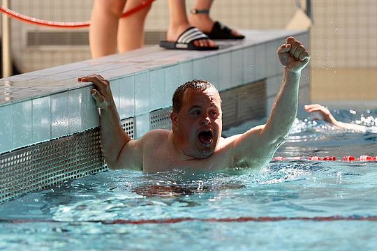 Ein Mann in einem Schwimmbad feiert mit erhobener Faust und hält sich am Beckenrand fest. Er sieht freudig und triumphierend aus. Im Hintergrund ist der Arm eines anderen Schwimmers zu sehen.
