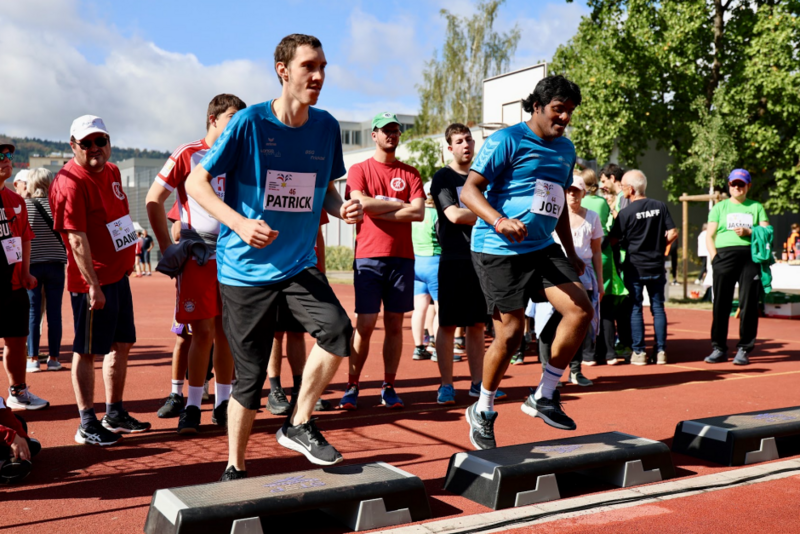 Zwei Männer in blauen Hemden betreten während einer Sportveranstaltung im Freien schwarze Podeste, während im Hintergrund an einem sonnigen Tag eine Gruppe von Menschen in Sportkleidung zusieht.