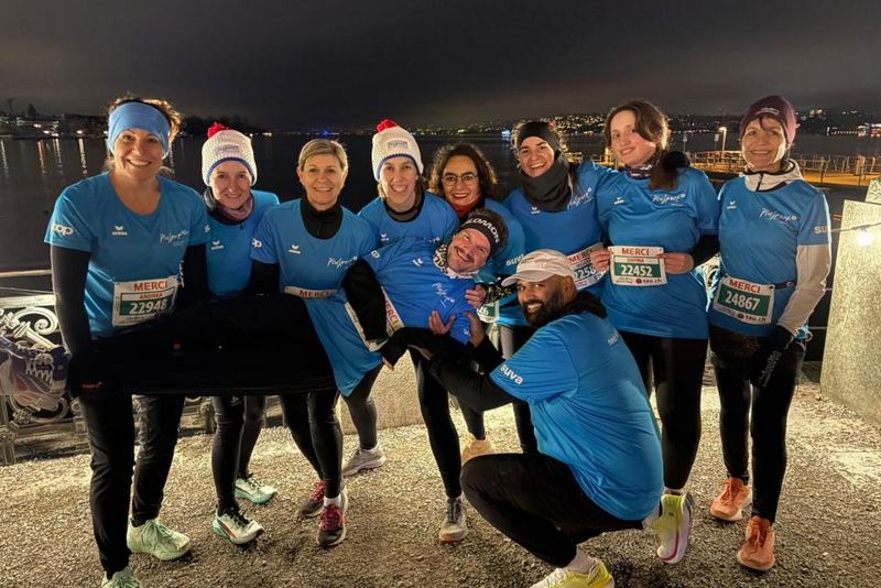 Eine Gruppe von elf lächelnden Läufern in passenden blauen Hemden posiert nachts zusammen und hält einen Teamkollegen waagerecht hoch. Sie tragen Startnummern und Wintermützen und stehen an einer Uferpromenade mit den Lichtern der Stadt im Hintergrund.