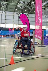 Eine Person im Rollstuhl spielt in der Halle Badminton – gerade beim Aufschlag – und trägt ein rotes Shirt und eine weiße Mütze. Im Hintergrund sind bunte Banner und Netze zu sehen, auf dem Spielfeld stehen orangefarbene Kegel.