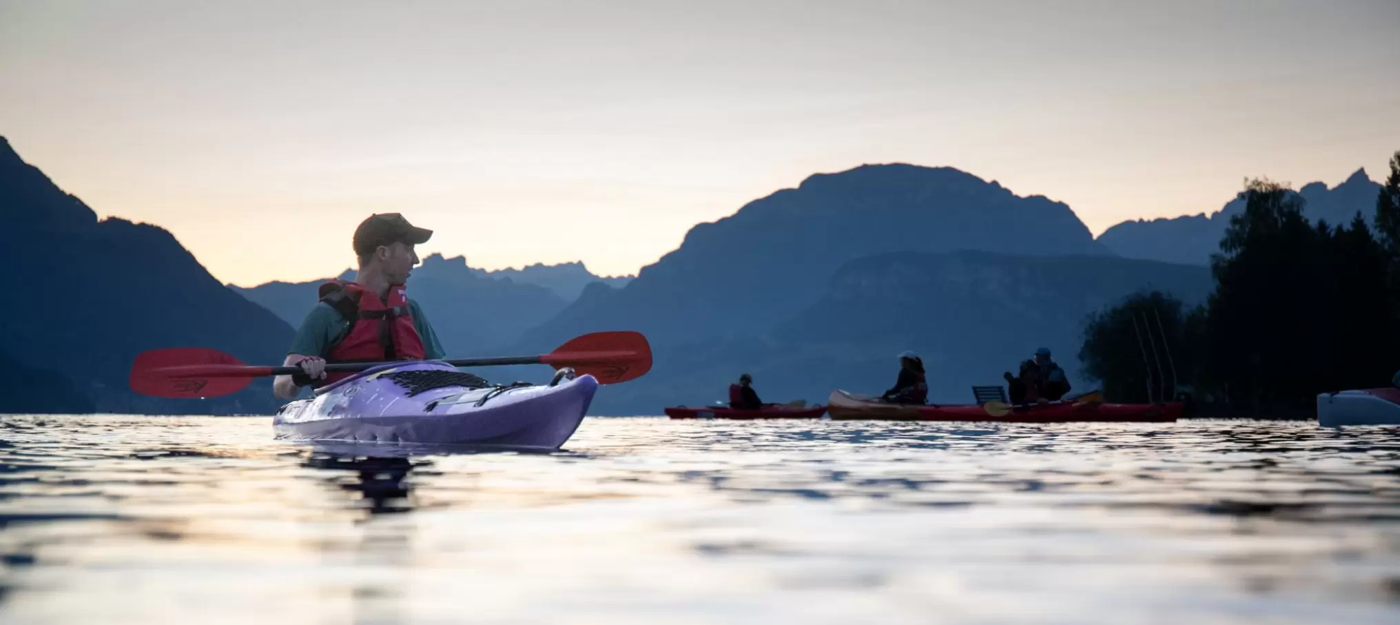 Eine Person in einem violetten Kajak paddelt in der Abenddämmerung auf einem See, im Hintergrund sind weitere Kajakfahrer zu sehen, und die Berge zeichnen sich als Silhouetten vor dem Abendhimmel ab.