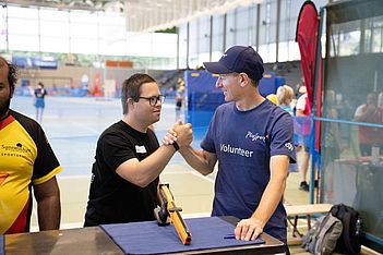 Zwei Männer geben sich bei einer Hallensportveranstaltung herzlich die Hand. Der eine trägt eine Brille und ein schwarzes T-Shirt, der andere ein blaues „Volunteer“-T-Shirt und eine Kappe. Im Hintergrund sind Sportgeräte und weitere Personen zu sehen.