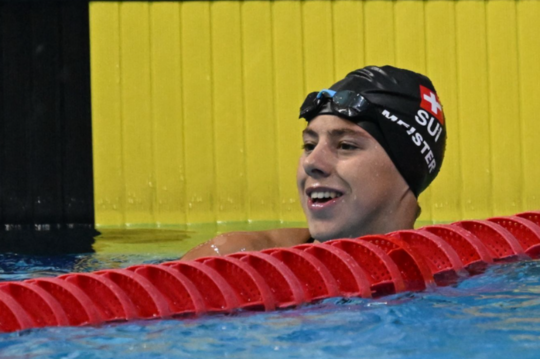 Ein Schwimmer mit schwarzer Badekappe und Schwimmbrille lächelt, während er am Beckenrand neben einer roten Bahnmarkierung und einer gelben Wand eine Pause macht.