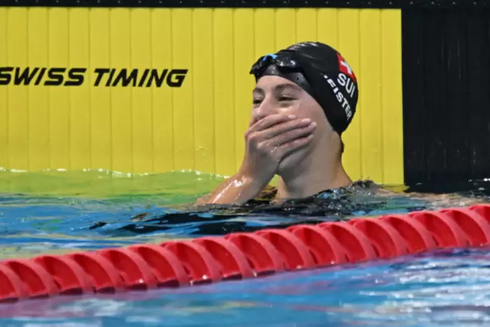Ein Schwimmer mit Badekappe und Schwimmbrille steht in einem Schwimmbecken, hält sich mit einer Hand den Mund zu und wirkt überrascht oder bewegt. Im Hintergrund sind eine rote Bahnbegrenzung und eine gelbe Wand mit der Aufschrift „SWISS TIMING“ zu sehen.