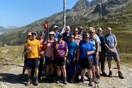 Eine Gruppe Wanderer steht gemeinsam auf einem Bergpfad und posiert für ein Foto vor einem hohen Wegweiser mit Flaggen. Im Hintergrund sind felsige Berge und grasbewachsene Hügel unter einem klaren blauen Himmel zu sehen.