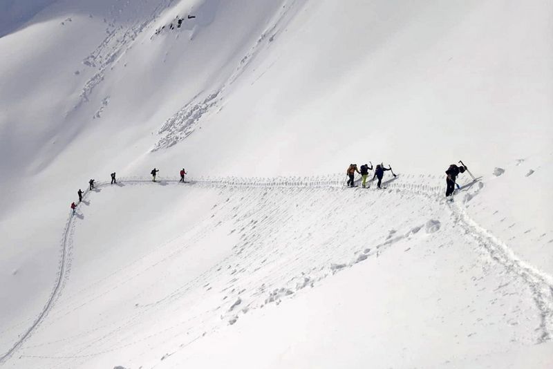 Eine Gruppe von Menschen in Winterkleidung wandert hintereinander einen schmalen, schneebedeckten Bergkamm entlang und hinterlässt unter einem blassen Himmel tiefe Fußspuren im Schnee.
