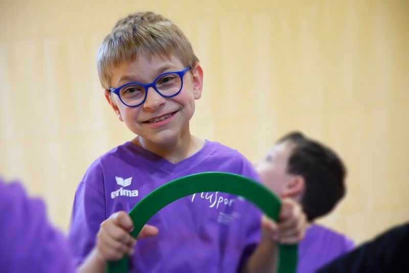 Ein lächelnder Junge mit Brille und lila Hemd hält einen grünen Reifen in der Hand. Ein anderes Kind, ebenfalls in lila, ist im Hintergrund zu sehen.