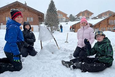 Vier Kinder in Winterkleidung sitzen und knien im Schnee mit einer kleinen Schneehütte und einer weißen Flagge. Im verschneiten Hintergrund sind Holzhäuser und weitere Menschen zu sehen, wobei der Schnee aktiv fällt.