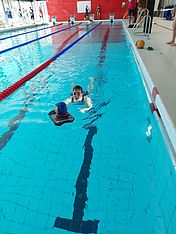 Eine Person schwimmt in einem Hallenbad auf ein im Wasser schwimmendes blaues Objekt zu. Andere Personen stehen am Beckenrand, und die Schwimmbahnen sind durch rote und blaue Seile voneinander getrennt.