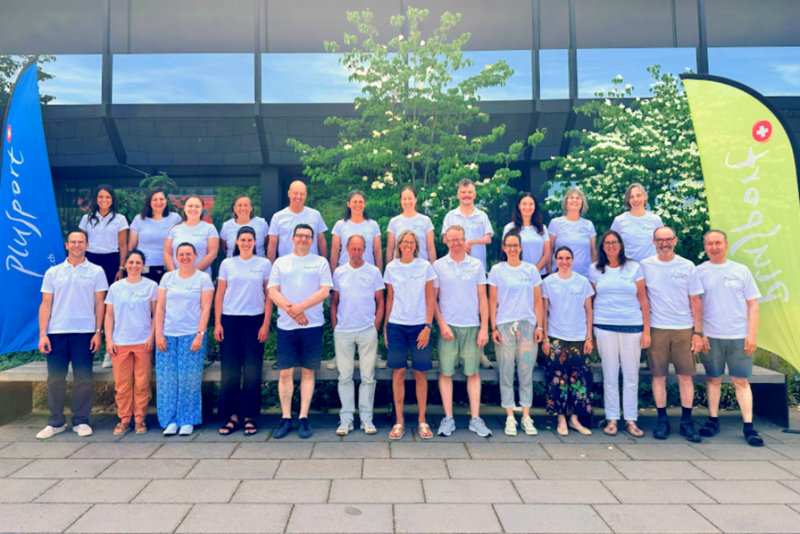 Eine Gruppe von 23 Personen in einheitlichen weißen Hemden steht im Freien vor einem Gebäude und grünen Bäumen und posiert für ein Gruppenfoto. Zu beiden Seiten der Gruppe sind blaue und grüne Fahnen mit Schriftzügen zu sehen.