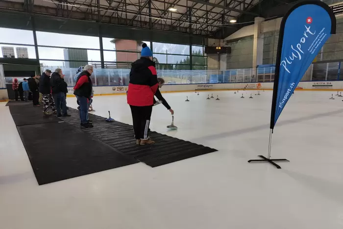 Menschen stehen auf einer schwarzen Matte in einer Eissporthalle und bereiten sich auf ein Spiel mit Curlingsteinen vor. Rechts steht eine blaue Flagge mit dem PluSport-Logo. Die Eisbahn ist von Brettern und Fenstern umgeben.