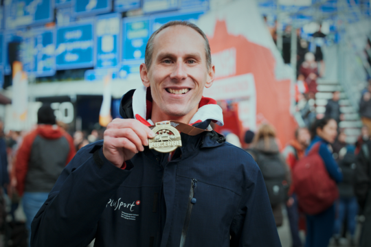 Ein lächelnder Mann in einer dunklen Jacke hält eine Goldmedaille vor einer belebten Szene im Freien mit Menschen und blauen Schildern im Hintergrund hoch.