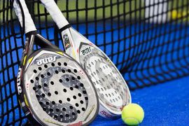 Zwei Padel-Schläger und ein gelber Tennisball liegen auf einem blauen Spielfeld neben einem schwarzen Netz. Die Schläger sind gegen das Netz gelehnt, ihre Vorderseiten zeigen kreisrunde Löcher.