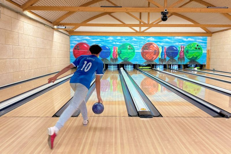 Eine Person in einem blauen Trikot spielt Bowling und ist dabei, eine blaue Bowlingkugel in eine Bahn einer hell erleuchteten Bowlingbahn zu werfen, an deren Rückwand bunte Bowlingkugel-Kunstwerke hängen.