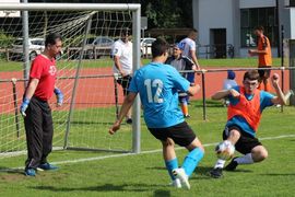 Ein Fußballtorwart in Rot sieht zu, wie ein Spieler in Blau (Nr. 12) einem anderen Spieler in Orange und Blau gegenübersteht, der in der Nähe des Tores rutscht und versucht, den Ball zu blocken oder zu schießen. Zuschauer und Spieler befinden sich im Hintergrund.