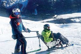 Eine Person in Skiausrüstung steuert einen Sitzski, auf dem eine andere Person sitzt, auf einem verschneiten Berghang, mit schneebedeckten Bergen und Bäumen im Hintergrund.