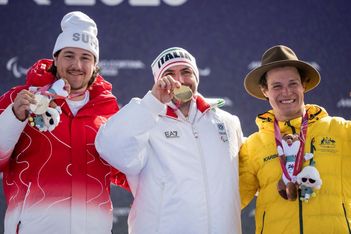 Drei männliche Sportler lächeln und zeigen ihre Medaillen bei einer Wintersportveranstaltung. Der Sportler in Rot hält eine Silbermedaille, der Sportler in Weiß eine Goldmedaille und der Sportler in Gelb eine Bronzemedaille.