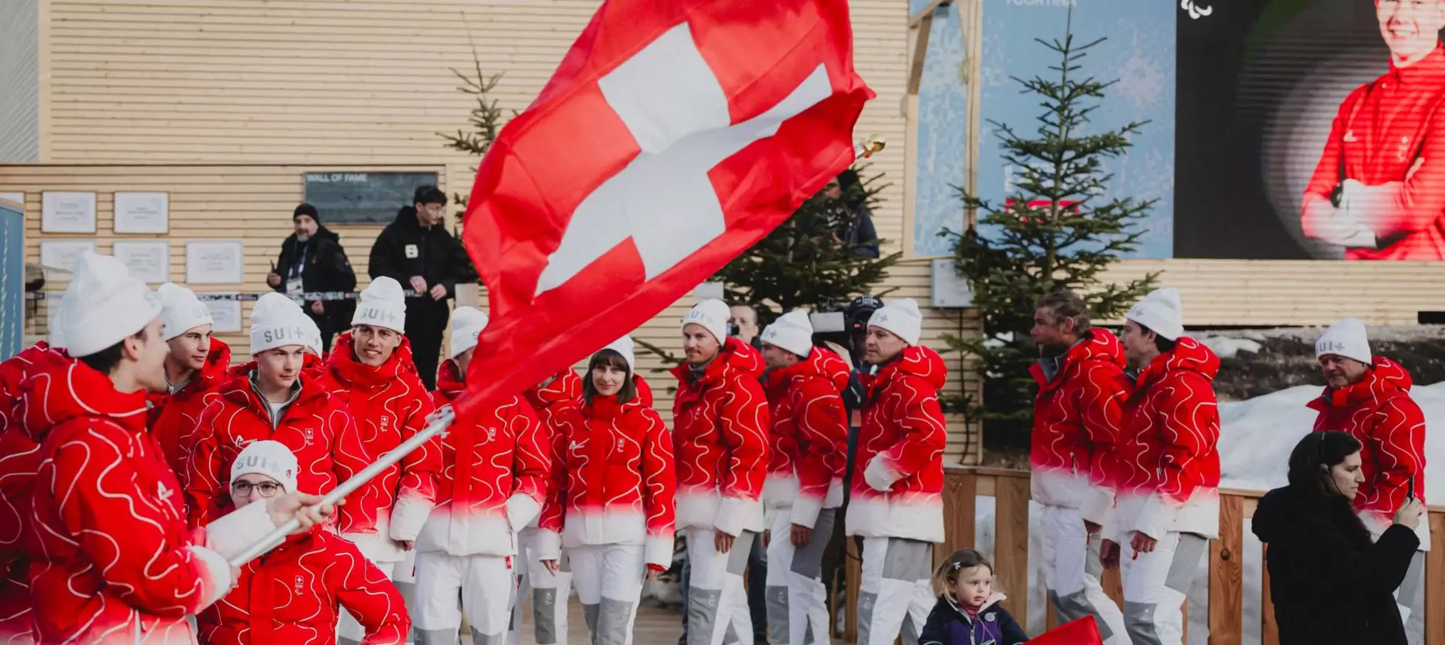 Eine Gruppe von Menschen in rot-weißer Kleidung steht zusammen, einer hält eine große Schweizer Flagge. Einige tragen Hüte mit der Aufschrift SUI. Im Hintergrund sind immergrüne Bäume und ein großer Bildschirm zu sehen. Vorne steht ein Kind.