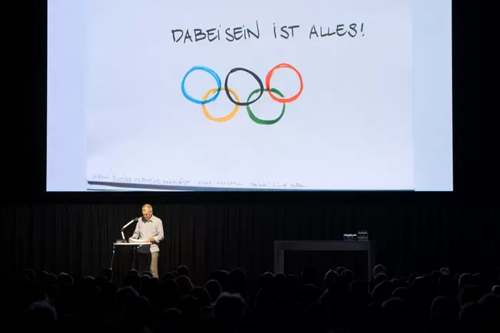 Eine Person steht auf einem Podium auf der Bühne und spricht zum Publikum. Dahinter ist eine große Leinwand mit den olympischen Ringen und dem deutschen Spruch „Dabeisein ist alles!“ zu sehen.