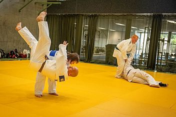 Zwei Paare trainieren Judo auf einer gelben Matte. Im Vordergrund wird eine Person geworfen. Im Hintergrund hält eine andere Person ihren Gegner am Boden fest. Alle tragen weiße Judo-Anzüge.