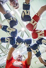 Eine Gruppe von Menschen, die Boxhandschuhe tragen, bildet einen Kreis und berührt sich in der Mitte mit den Handschuhen – aus der Untersicht gefilmt. Zwei Handschuhe sind rot, die übrigen sind blau. Die Gesichter sind zu sehen, wie sie zur Kamera hinunterblicken.