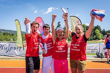 Vier Menschen in roten Trikots feiern auf einer Sportbahn, halten Wasserflaschen in den Händen und strecken die Arme in die Höhe, im Hintergrund sind an einem sonnigen Tag bunte Fahnen und Bäume zu sehen.