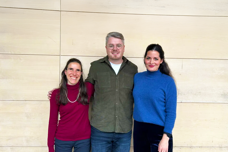 Drei Erwachsene stehen lächelnd vor einer hellen Holzwand. Die Person links trägt einen roten Rollkragenpullover, die Person in der Mitte eine Brille und ein grünes Hemd und die Person rechts einen blauen Pullover.