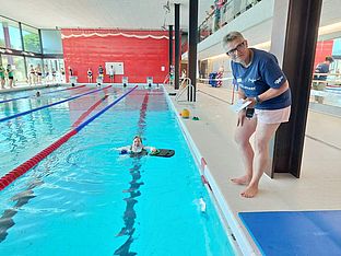 Eine Person steht am Beckenrand und beobachtet einen Schwimmer, der in einem Hallenbad ein Kickboard benutzt. Im Hintergrund sind weitere Schwimmer und Menschen zu sehen.