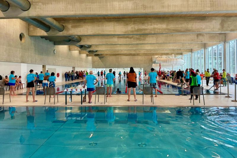 Ein Schwimmwettkampf in einem Hallenbad mit Schwimmern, Trainern und Zuschauern. Die Leute stehen am Beckenrand, während andere hinter Absperrungen zuschauen. Der Schwimmbereich hat große Fenster und Betonbalken über dem Kopf.